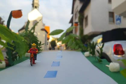 Im Vordergrund fährt ein Lego-Männchen mit einem Lego-Fahrrad auf einer gebastelten Straße aus Pappe während im Hintergrund eine echte Straße zu sehen ist