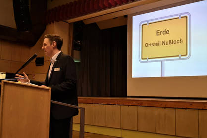 Mann am Rednerpult am Sprechen, im Hintergrund eine Präsentationsfolie, die ein Ortschild zeigt mit der Aufschrit "Erde, Ortsteil Nußloch"